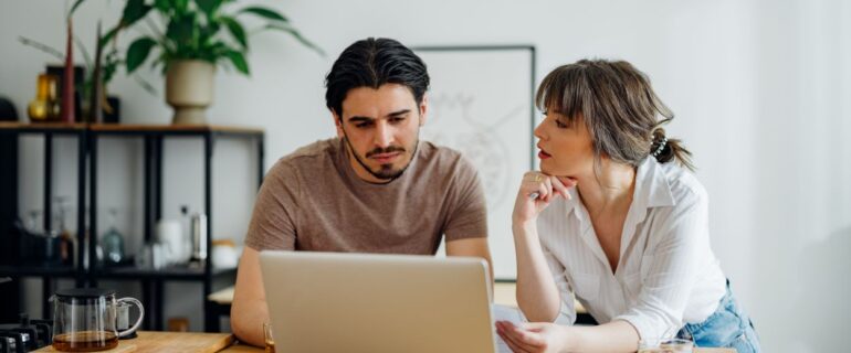 Boyfriend and girlfriend talking and doing home finances together online on a laptop computer in the kitchen.