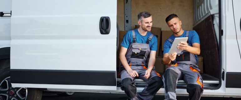 Two men look at a tablet while sitting on the kickboard of a white van